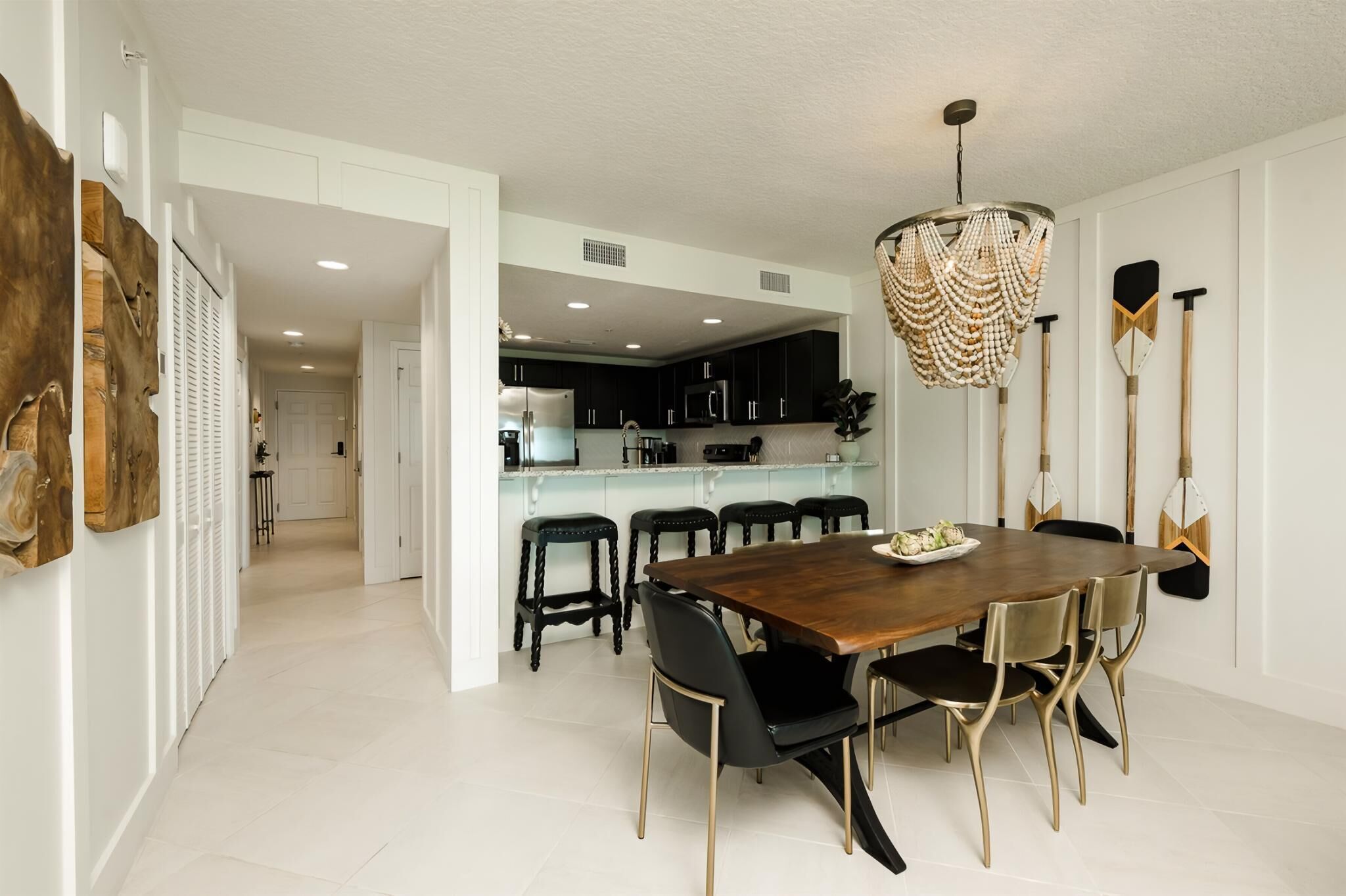 A modern dining area with a wooden table and black chairs, a chandelier, and a kitchen in the background.