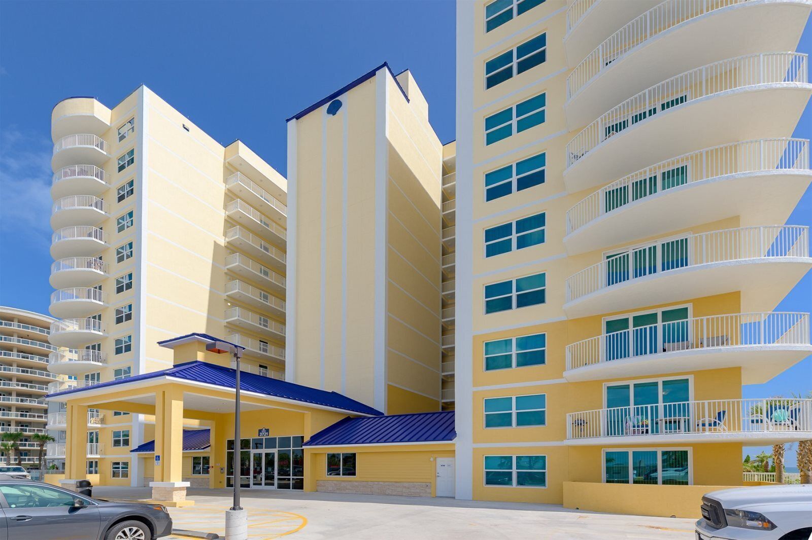 Un hotel de varios pisos con balcones, un toldo azul y un estacionamiento.