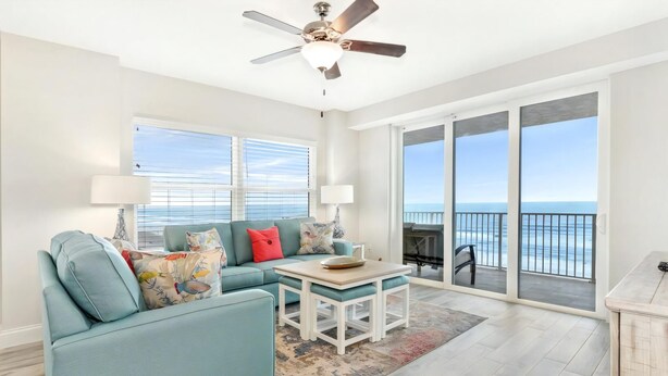 A spacious living room with a blue sofa, a coffee table, and a ceiling fan. Large windows offer a view of the ocean. - Aruba Collection (Daytona Beach Shores)