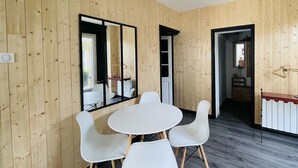 Una habitación con paredes de madera, una mesa redonda blanca con cuatro sillas, una lámpara en el techo y una puerta que da a otra habitación.