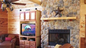 A cozy cabin interior with a stone fireplace, a television mounted on a wooden cabinet, and leather furniture.