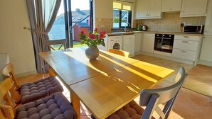Un coin repas avec une table en bois et des chaises rembourrées, une cuisine en arrière-plan et une vue sur l’extérieur.