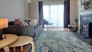 Un salon comprenant un canapé, une table basse, un tapis, une cheminée et une vue sur l’extérieur par une fenêtre ornée de rideaux.