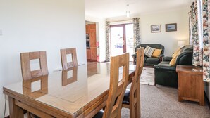 A dining area with a glass-top table and wooden chairs, a green sofa with cushions, and a small wooden side table. - Woodburn Cottage (Waternish)