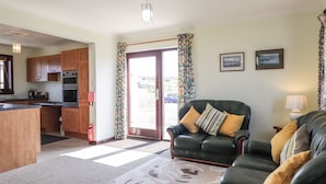 A living room with a sofa and armchairs, a kitchen area with wooden cabinets, and a large window with curtains. - Woodburn Cottage (Waternish)