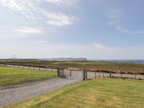 A rural landscape with a gravel path leading to a metal gate, grassy fields, and distant hills. - Woodburn Cottage (Waternish)