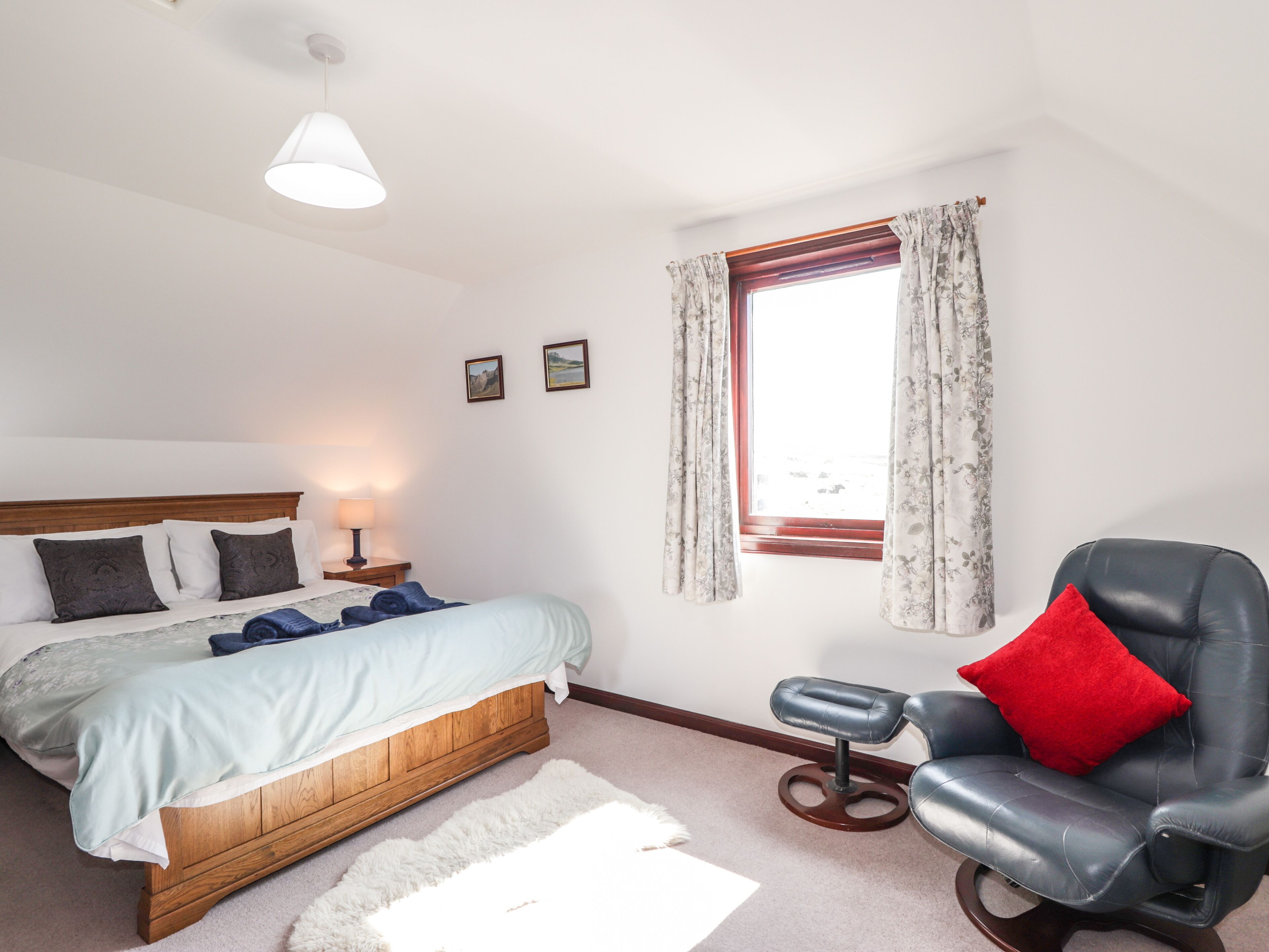 Een slaapkamer met een bed, een stoel, een raam met gordijnen, een lamp en een klein krukje.