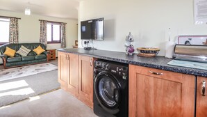 A compact living space with a kitchen area, a washing machine, a flat-screen TV, and a sofa with cushions. - Woodburn Cottage (Waternish)