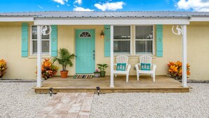 Uma casa com varanda, duas cadeiras brancas, uma porta verde e um pequeno jardim.