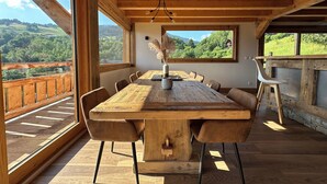 A wooden dining table with chairs, a vase with dried flowers, and a view of a green landscape through large windows. - Chalet Mont Blanc view with outdoor jacuzzi (Combloux)