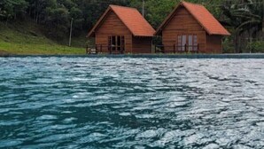 Two wooden cabins with red roofs, situated on a lakeside with a forested hill in the background. - Pousada Rio Túnel (Grão Pará)