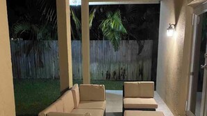 Een terras buiten met een loungeset en een rieten poef, een plafondventilator en een houten omheining op de achtergrond.