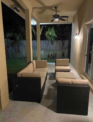 An outdoor patio with a sofa set and a wicker ottoman, a ceiling fan, and a wooden fence in the background.