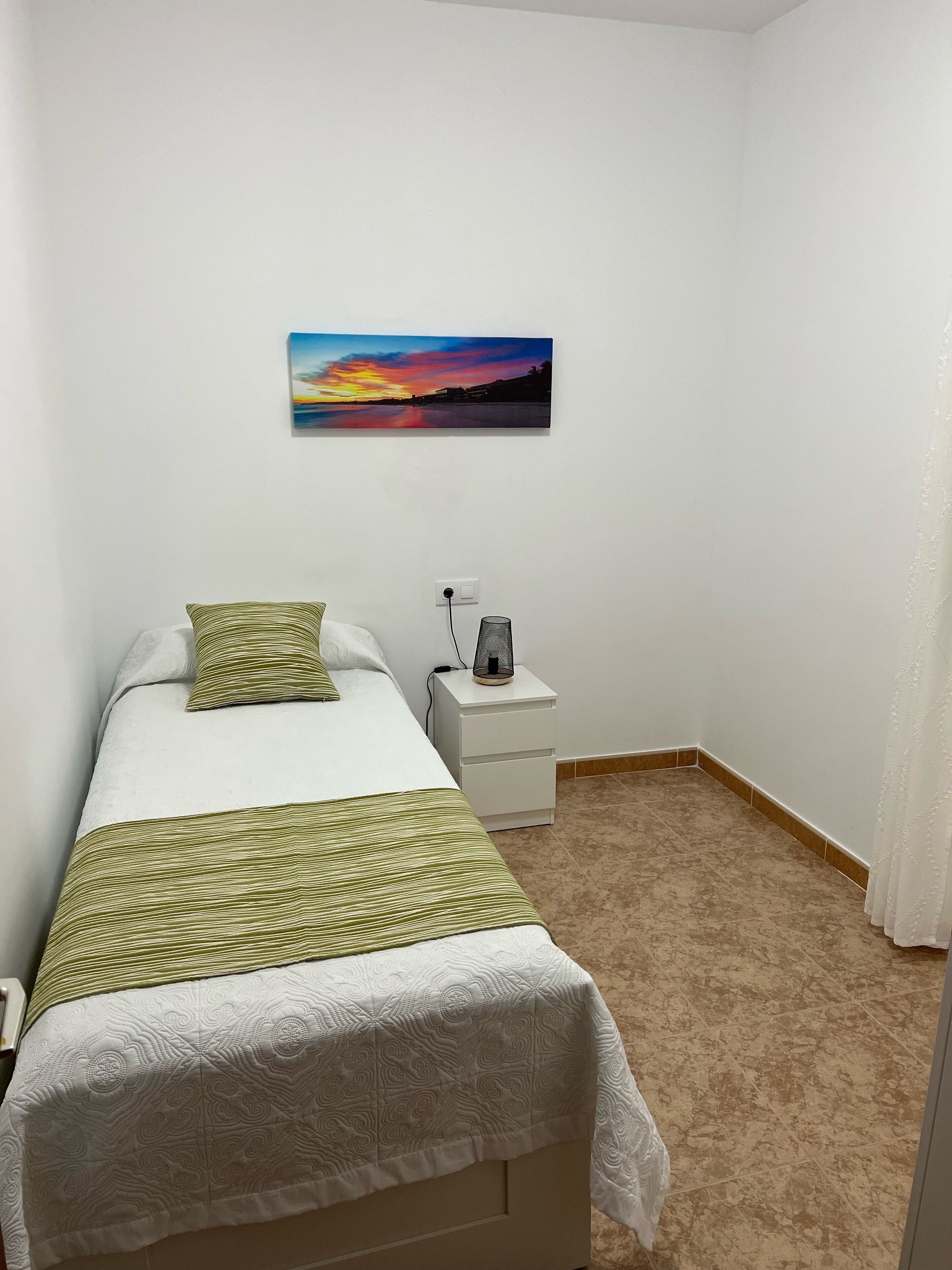 A bedroom with a bed, a nightstand, a power outlet, and a painting on the wall.
