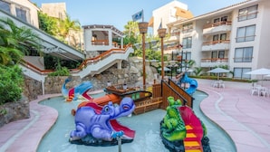 Ein Wasserpark mit Rutschen in Form eines Elefanten und eines Krokodils, umgeben von einem Pool und einem Gebäude mit Balkonen.