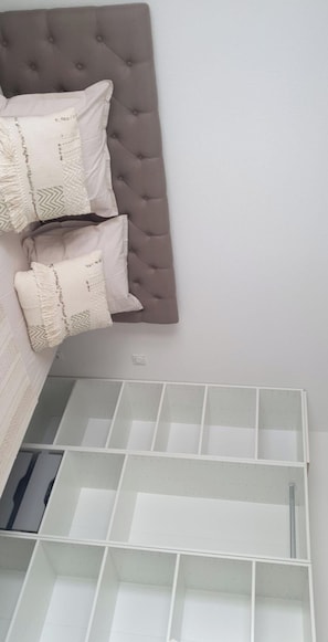 Quarto com cabeceira estofada, duas almofadas estampadas e um móvel de estante branco abaixo.