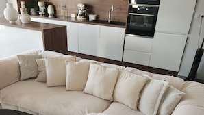 Una sala de estar moderna con un sofá seccional color beige, un tapete blanco mullido y una zona de cocina con armarios y electrodomésticos blancos.