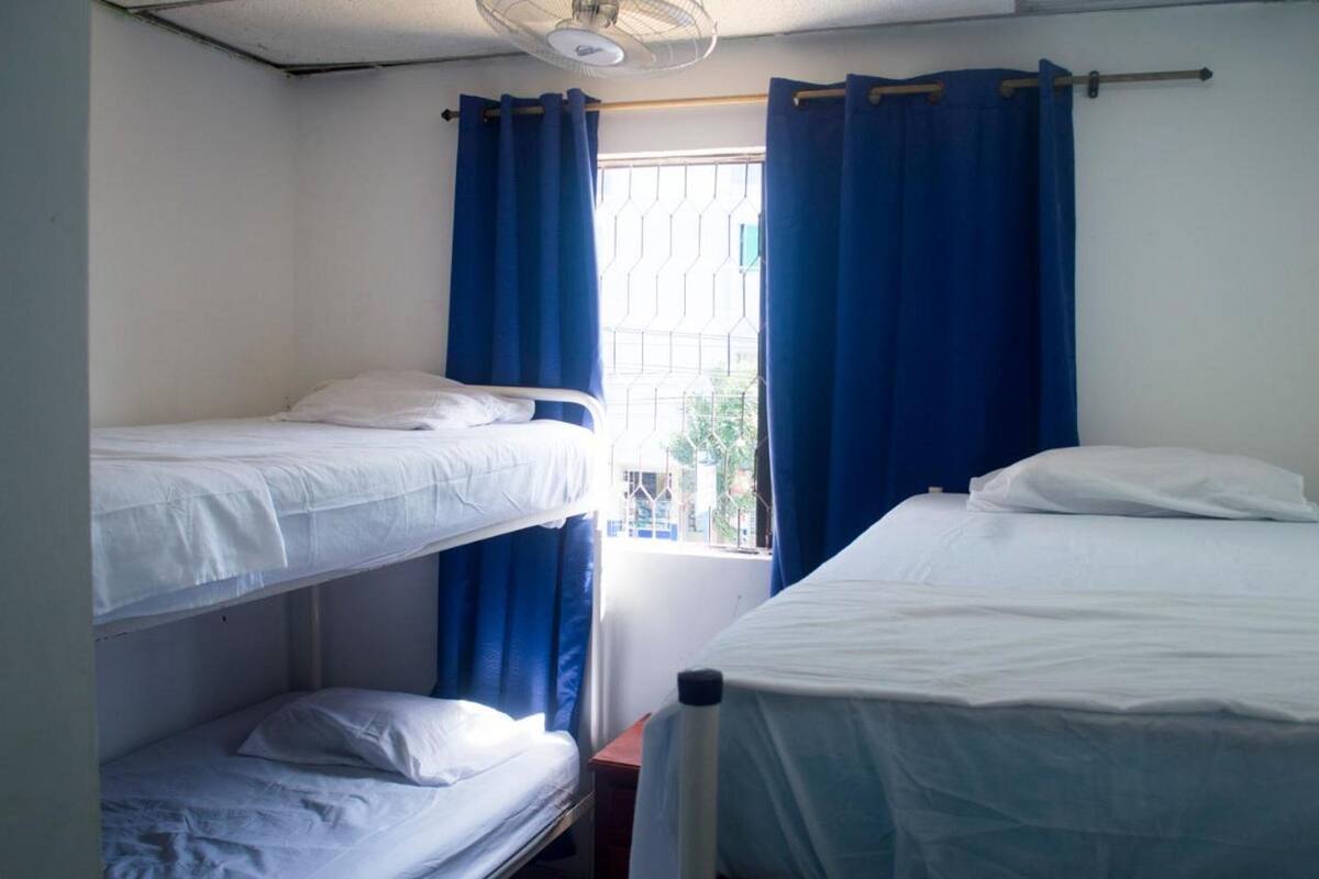 A dormitory room with bunk beds, a window with blue curtains, and a ceiling fan.