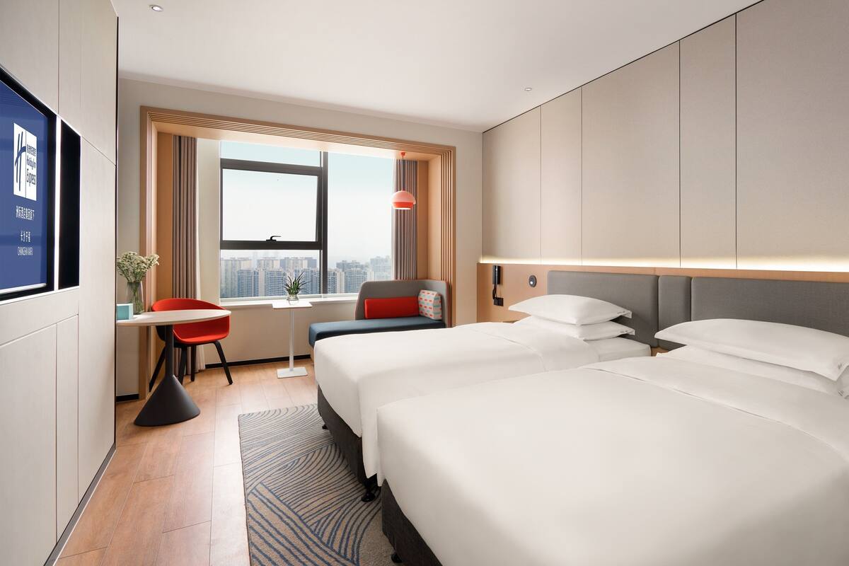 A hotel room with two beds, a desk, a chair, a TV, and a window with a city view.