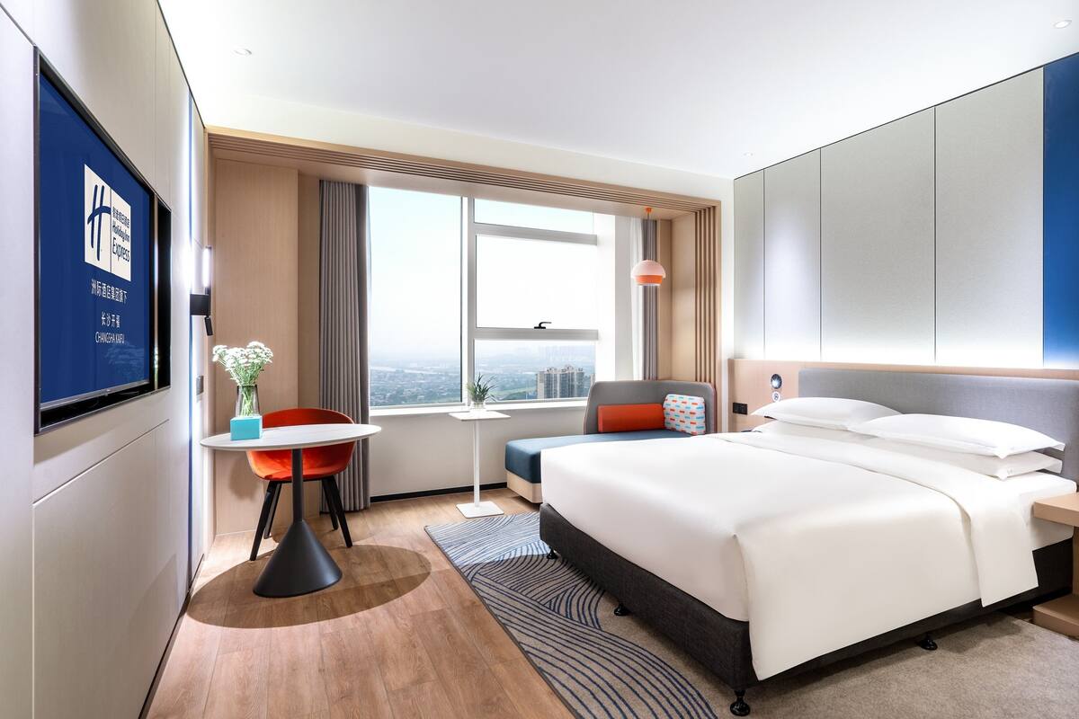 A hotel room with a bed, a desk, a chair, a TV, and a window with a city view.