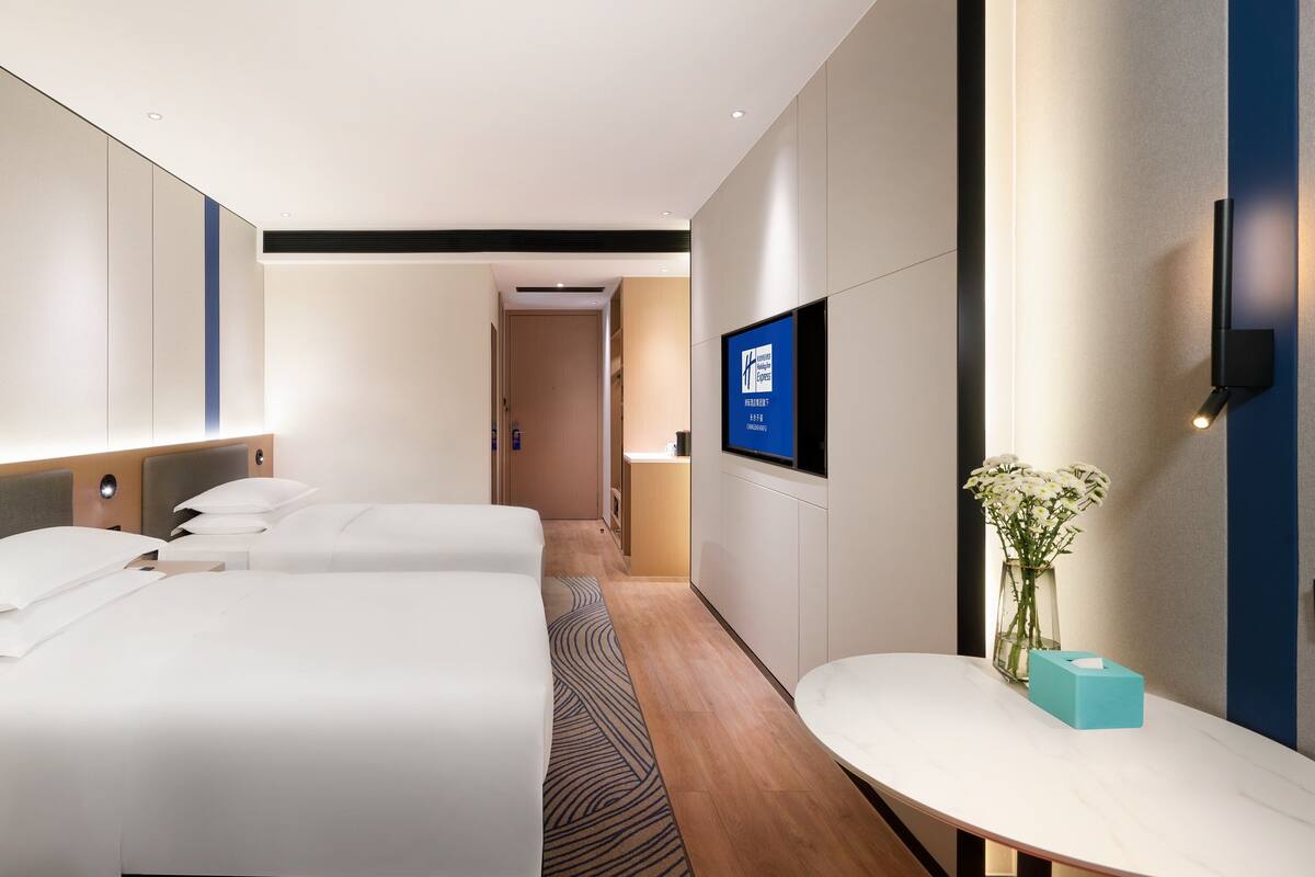 A hotel room with two beds, a small table, a TV, and a wall-mounted light.