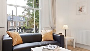 Ett modernt vardagsrum med en svart lädersoffa, ett vitt soffbord med en bok, ett litet vitt sidobord med en lampa och ett stort fönster med utsikt över en byggnad och träd.