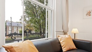 Ett rum med en svart lädersoffa, två guldfärgade kuddar, en lampa och utsikt över en gata med träd och byggnader.