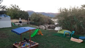 Ein Garten mit Spielbereich, darunter ein Spielhaus aus Holz, eine Rutsche und ein Bällebad. In der Nähe eines Gewässers stehen Stühle und ein Tisch, im Hintergrund Bäume und Berge.