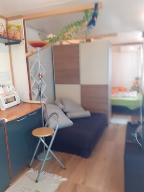 A compact living space with a kitchenette, a foldable chair, a bed with a dark cover, and a small table with a toaster.