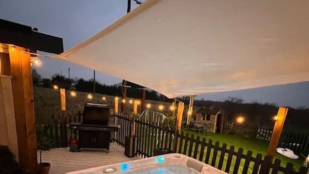 A hot tub on a wooden deck with string lights and a barbecue grill in the background.
