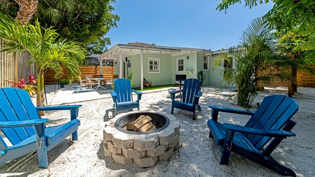 A patio area with blue Adirondack chairs, a stone fire pit, and a house in the background. - The Ivanhoe Hideaway (Sarasota)