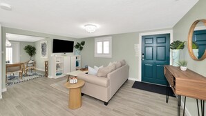 A modern living room with a beige sofa, wooden coffee table, and a TV on a white cabinet. A teal door and a mirror are visible. - The Ivanhoe Hideaway (Sarasota)
