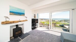 Un salon moderne avec une cheminée, un tableau représentant une plage et un balcon avec vue sur la mer.