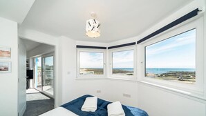 Une chambre à coucher avec un lit, deux fenêtres donnant sur la plage, un luminaire au plafond et une porte menant à un balcon.