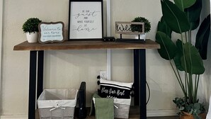 Una TV a schermo piatto montata a parete mostra un paesaggio suggestivo. Sotto la TV, un ripiano di legno con un cartello di benvenuto incorniciato, una piccola pianta in vaso e un oggetto decorativo.