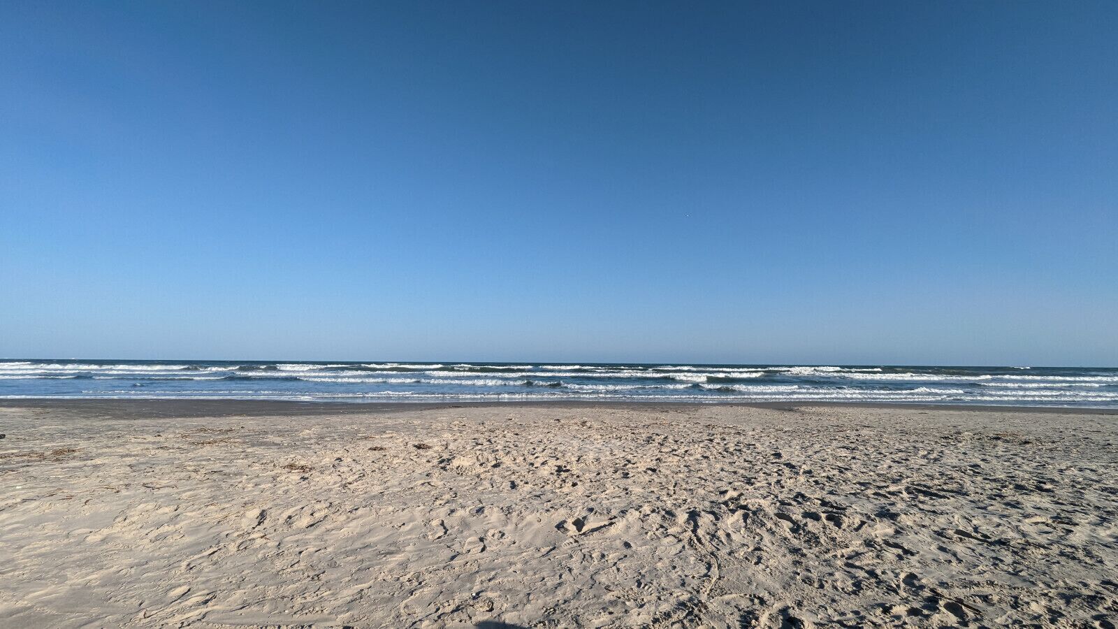 Une vaste plage de sable où les vagues se brisent près du rivage, sous un ciel dégagé.