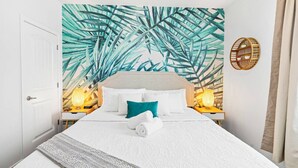 A hotel room with a bed, white walls, a tropical leaf mural, two bedside lamps, a round wooden shelf, and a white door.