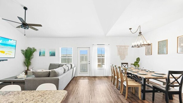 A modern, open-plan living and dining area with a wooden floor, a grey sofa, a dining table set for six, and a ceiling fan.