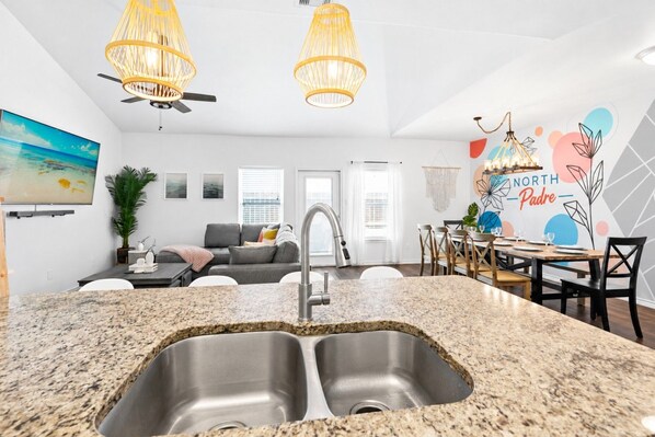 Una cocina moderna con encimera de granito, pileta de acero inoxidable y comedor con mesa y sillas de madera.