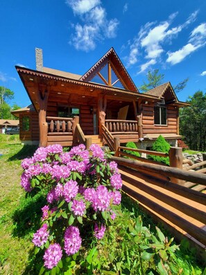 A wooden cabin with a porch and a garden with pink flowers. - Newly Built Handcrafted Cozy Log Home (Monticello)