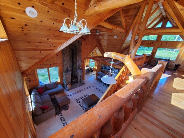 A spacious cabin with a stone fireplace, wooden beams, and large windows offering views of the outdoors. - Newly Built Handcrafted Cozy Log Home (Monticello)