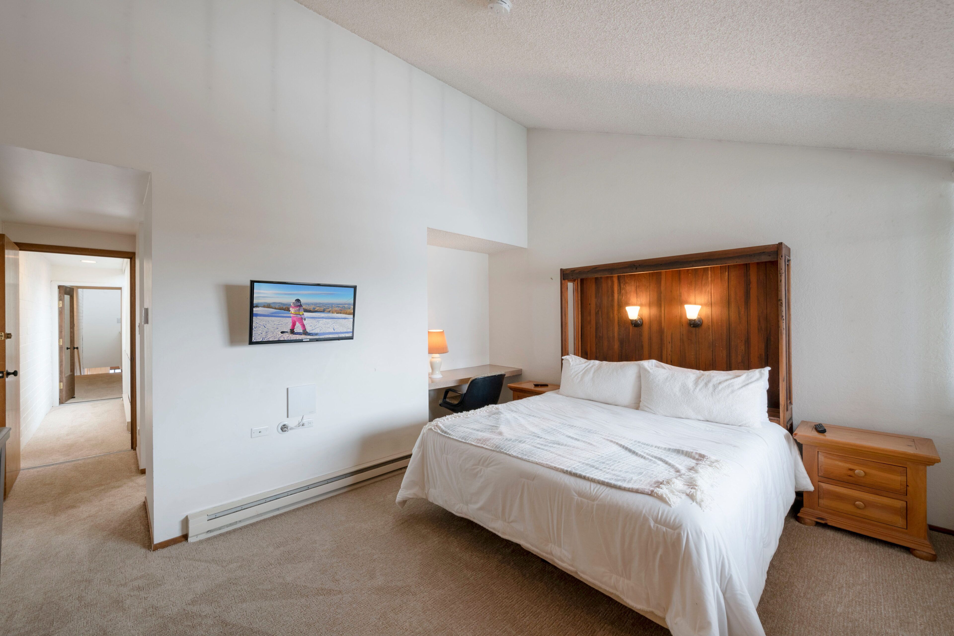 A bedroom with a large bed, a wooden headboard, a bedside table, a desk with a chair, and a wall-mounted TV.