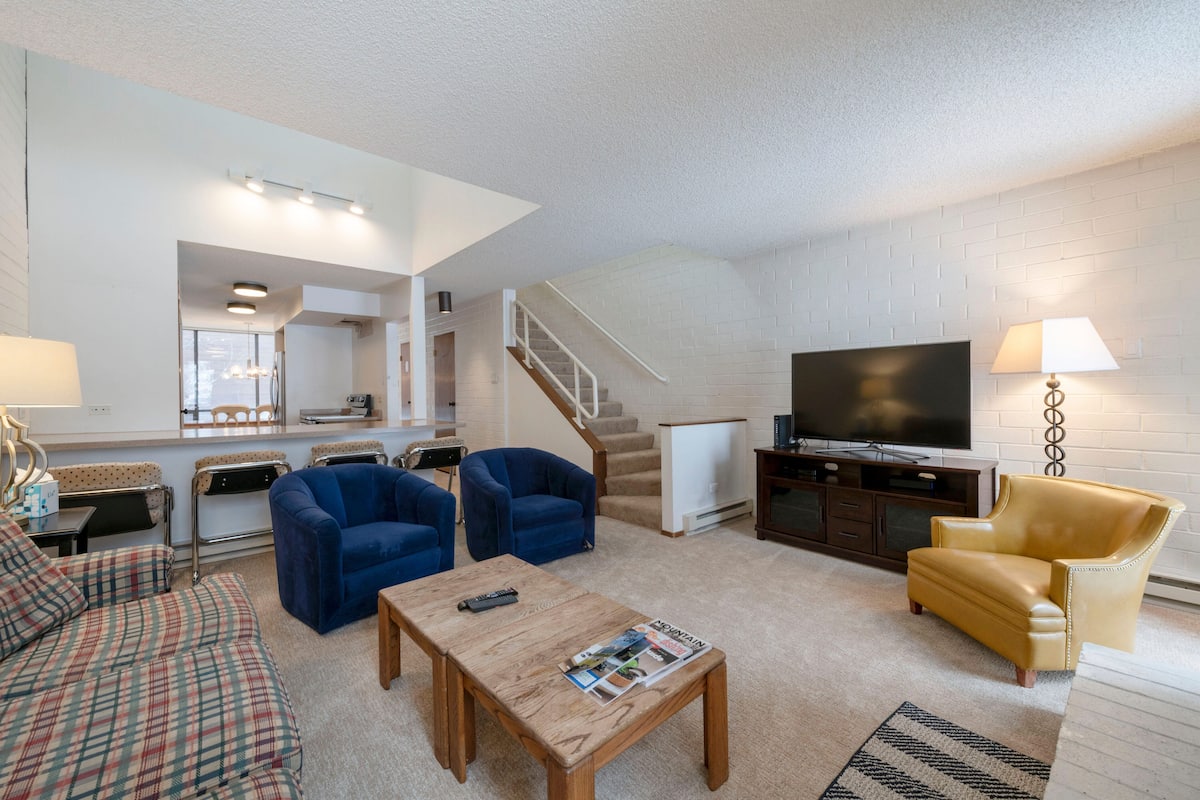 Ein Wohnzimmer mit einer Couch, zwei Sesseln, einem Couchtisch, einem Fernseher und einer Treppe, die zu einer oberen Ebene führt.