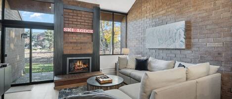 A modern living room with a fireplace, a sofa set, and a large window offering a view of the outdoors.