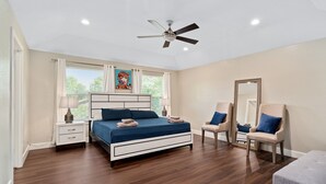 Quarto com cama, mesas de cabeceira, uma cadeira, um banco, ventoinha no teto e janela com cortinas.