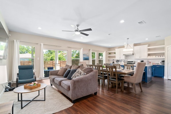A spacious living area with a sofa, coffee table, and dining table. There is a ceiling fan and a kitchen area with cabinets and a blue island.