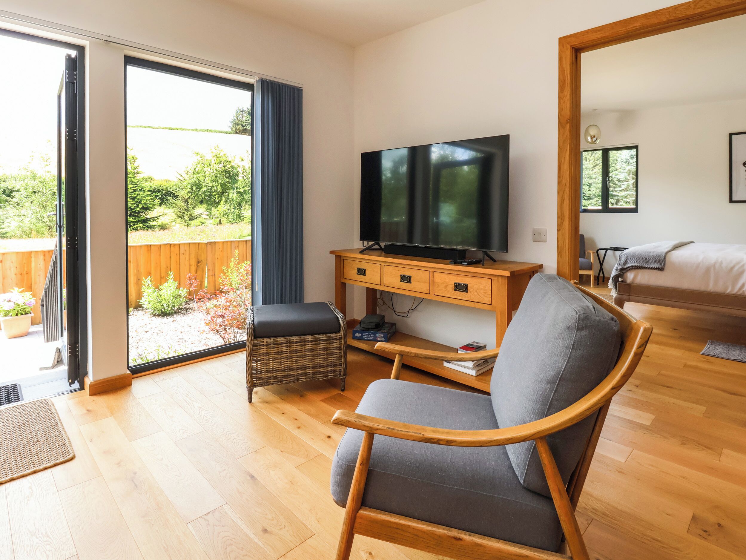 A modern living room with a wooden floor, a grey sofa, a wicker chair, a TV on a wooden stand, and a sliding glass door leading to an outdoor area with a wooden fence and plants.