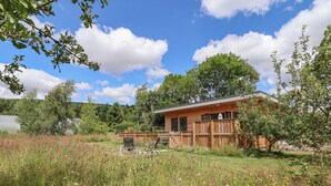 A wooden cabin with a deck, surrounded by a grassy field with wildflowers and trees. - No1 The Lodge (Bishops Castle)