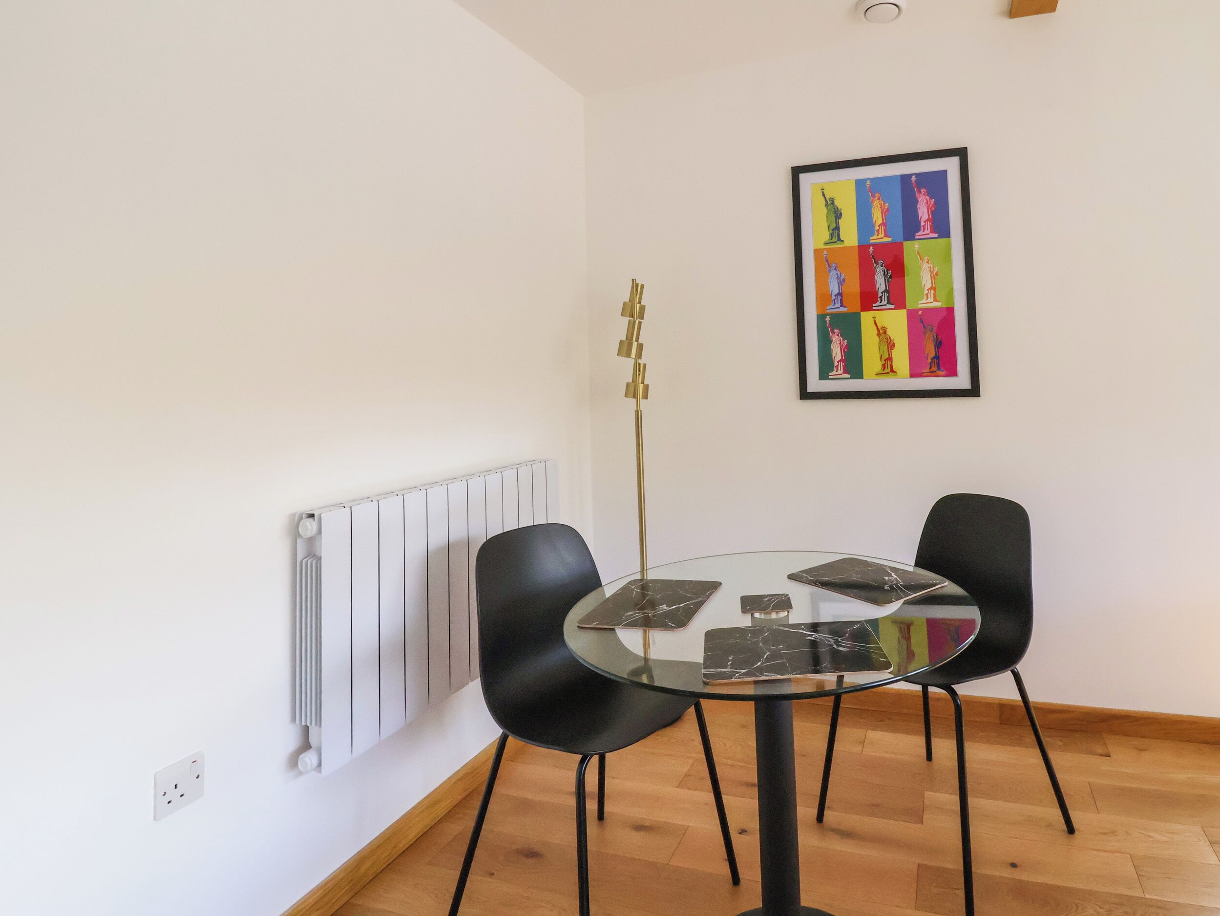 A dining area with a glass table and two black chairs, a radiator, and a colorful framed artwork on the wall.