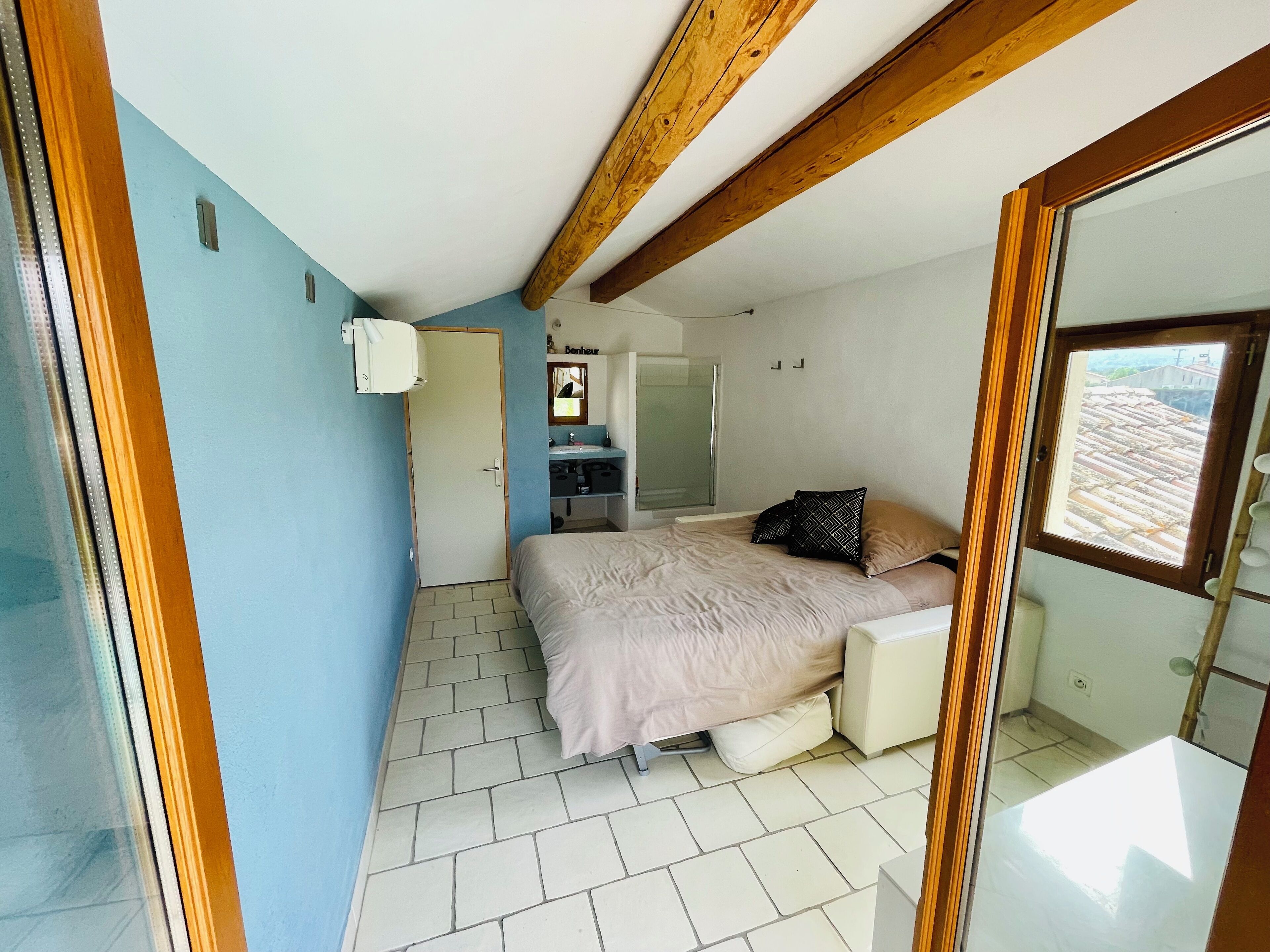 A bedroom with a bed, a small bedside table, a mirror, a window, and a bathroom with a sink and mirror.